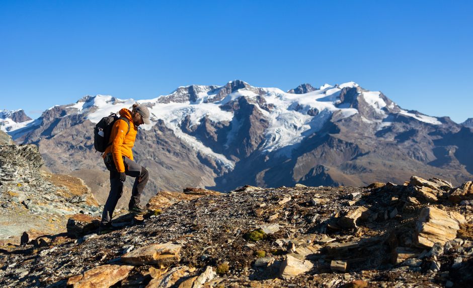 A hiker explores rocky trails in Staffal, Aosta Valley, with snow-capped Alps in the background.