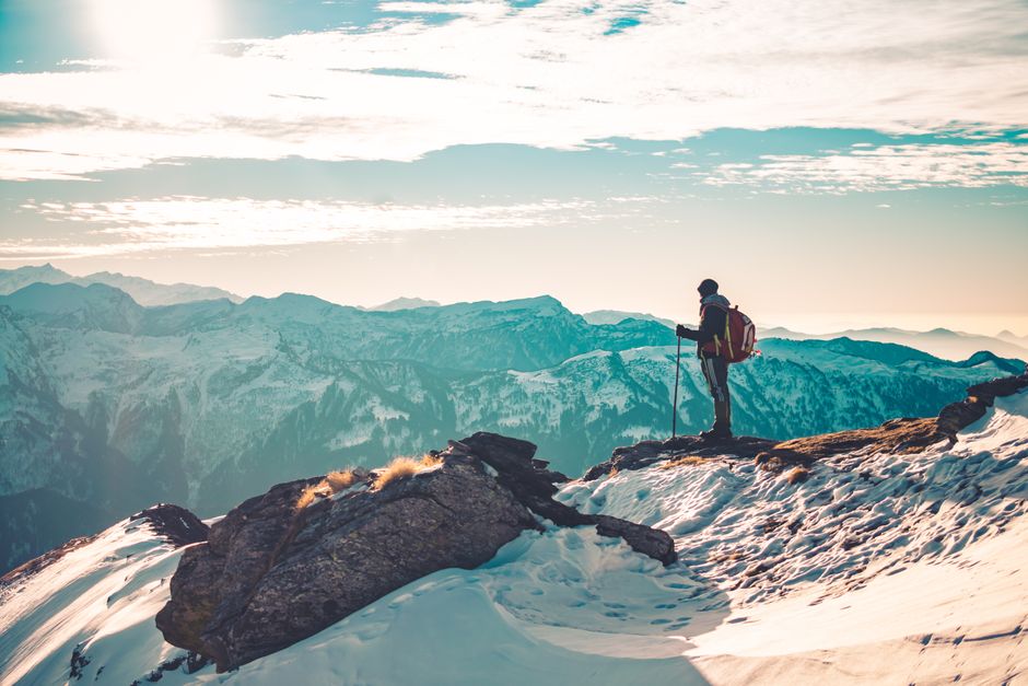 A lone hiker with trekking poles stands on a snowy mountain peak at sunrise.